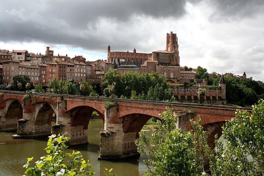 Una vista de Albi al otro lado del río que muestra un viejo puente de ladrillo rojo sobre el río, con casas antiguas que conducen a la catedral y el espectacular cielo nublado