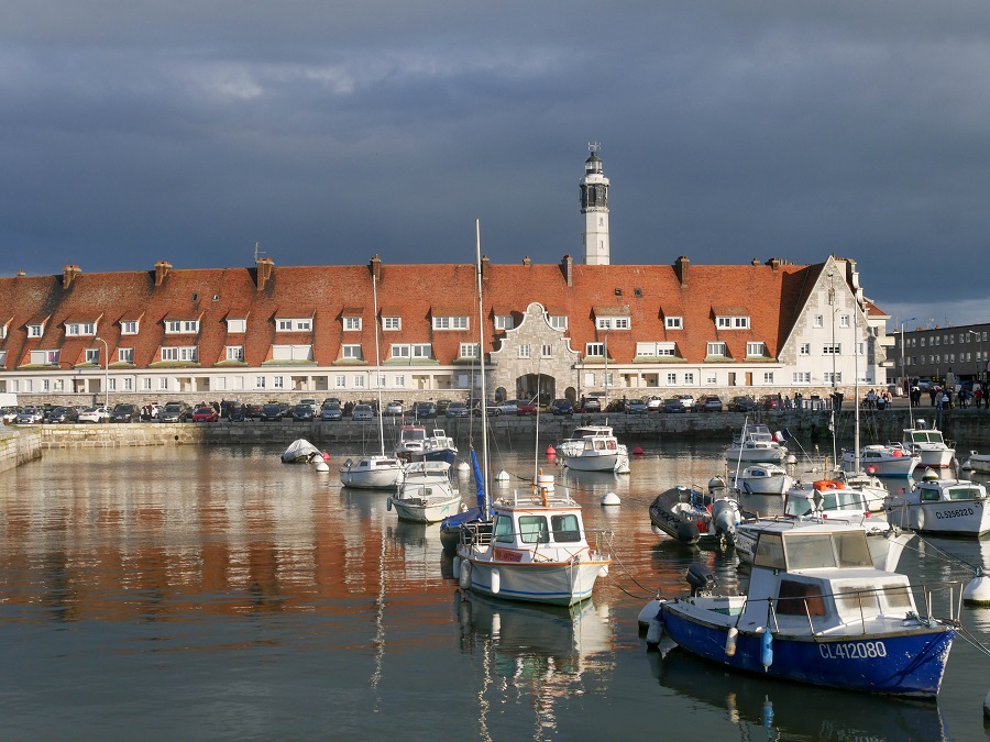 Calais Bay con botes flotando en el agua y un largo edificio detrás con techos rojos y un faro en la distancia