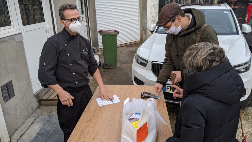 Comida para llevar con la historia de Ancienne con una mesa afuera en el estacionamiento, un chef detrás de la mesa, dos personas con una bolsa de comida y pagando una factura de tarjeta de crédito