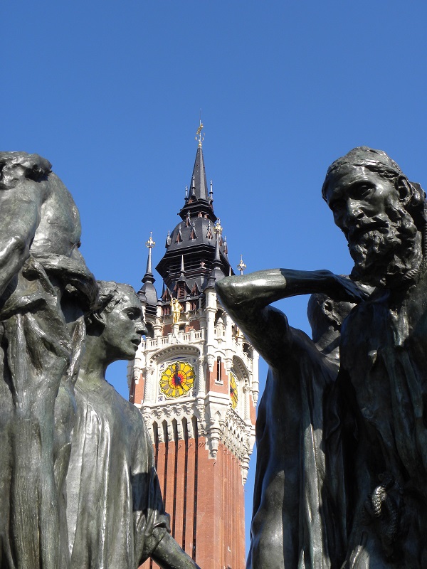 Primer plano de 2 tomas de los burgueses de Calais de Rodin con un alto campanario de ladrillo rojo en el fondo