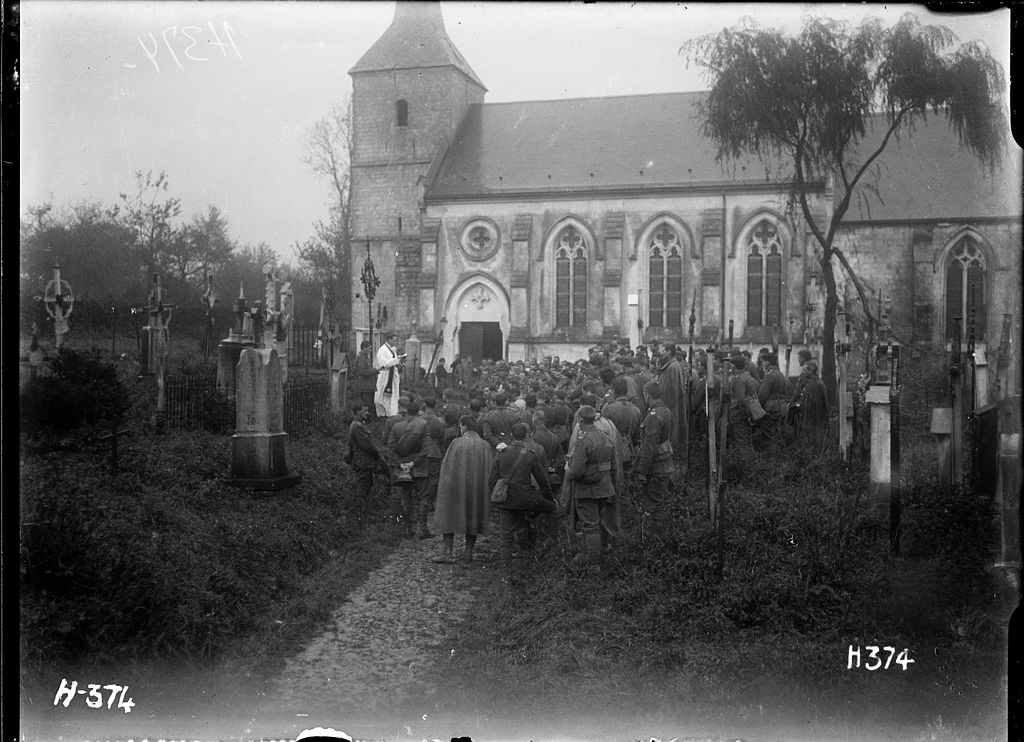 Fotografía en blanco y negro del Día de los Difuntos de 1917 en Pas de Calais con soldados neozelandeses reunidos alrededor de una tumba y un sacerdote fuera de una iglesia