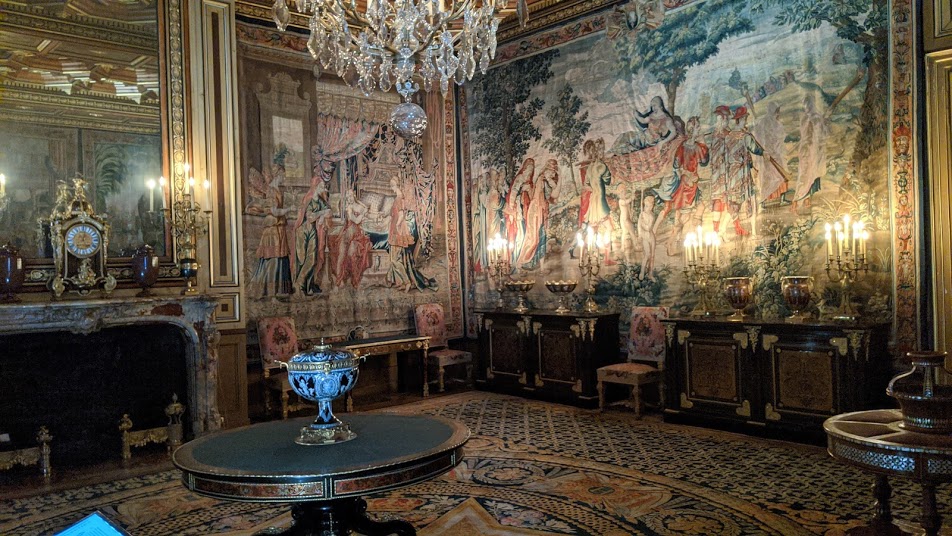 Salón de tapices en Fontainebleaus que muestra una gran sala iluminada por luces parpadeantes con dos paredes cubiertas con tapices que representan escenas de caza.