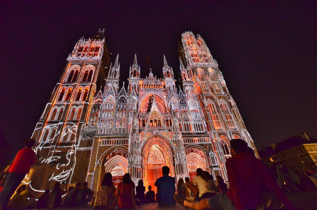 Catedral de Rouen con un colorido espectáculo de luces en la fachada. Mirando hacia arriba con gente directamente al frente y principalmente luces rojas y naranjas