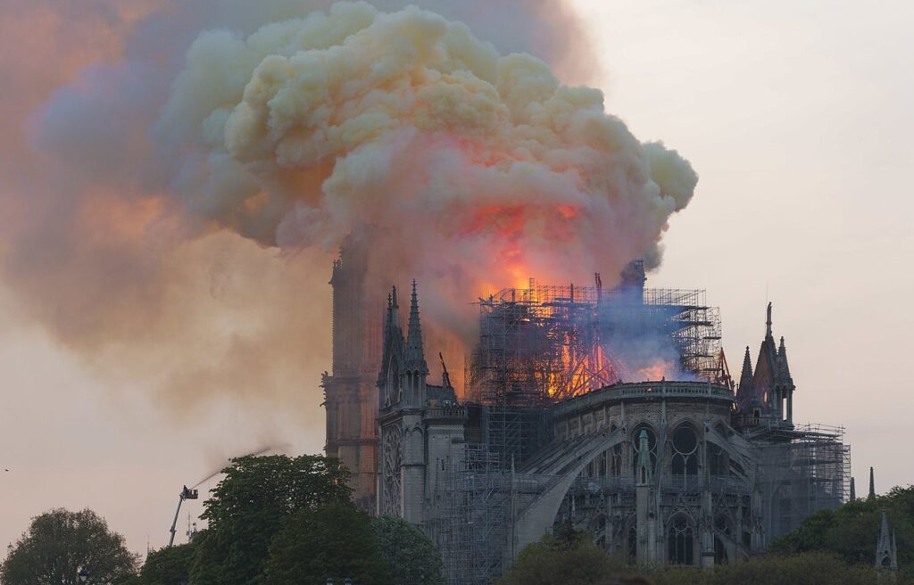 Notre-Dame de París en llamas con humo y llamas del techo tomadas desde atrás varias personas viendo el fuego