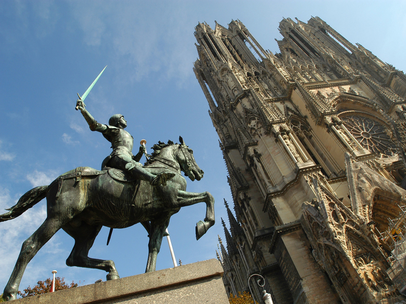 Mirando hacia arriba desde el suelo a la estatua de Juana de Arco sobre un pedestal. Ella está montando un caballo al galope con una espada en su mano derecha mirando directamente a la fachada de la catedral de Reims.