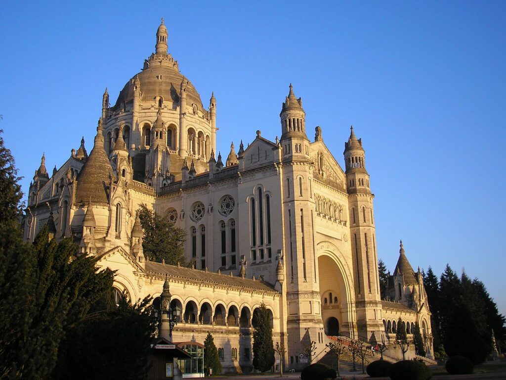 Exterior de la Basílica de Santa Teresa en Lisieux que muestra una enorme basílica con una nave larga en el frente y una pequeña torre detrás en la cálida luz del sol de verano