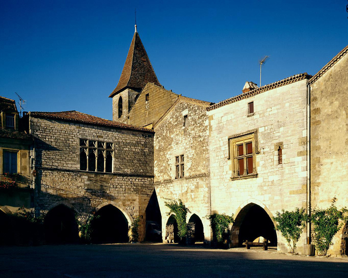 Monpazier, una bastida que muestra una esquina con edificios modestos y una iglesia detrás y arcadas cubiertas que recorren la plaza del mercado