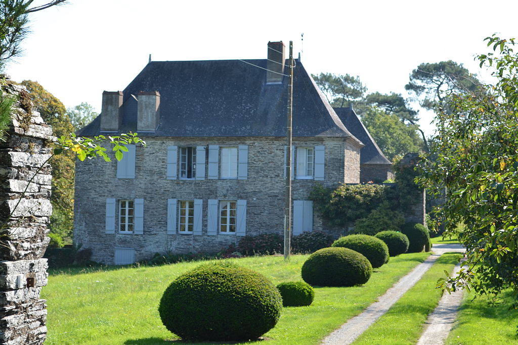 Maison de maitre en Loire Atlantique que muestra una gran casa de piedra de dos plantas con un techo inclinado inclinado, persianas en las ventanas que dan al césped en una pendiente con bojes cónicos de forma simétrica que bordean el camino.