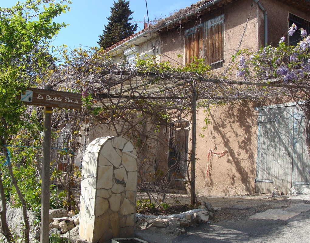 Bastide Neuve Marcel Pagnol, una bastida en el sur de Francia que muestra una gran piedra en el frente y una casa rosa de dos pisos con techo de tejas detrás en ángulo y enrejados que ocultan parte del frente