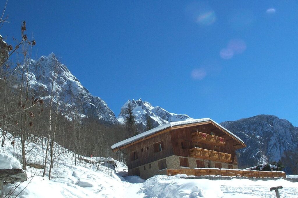 Chalet de madera moderno e independiente en los Alpes con nieve en el suelo y techo inclinado con una montaña y árboles en el fondo