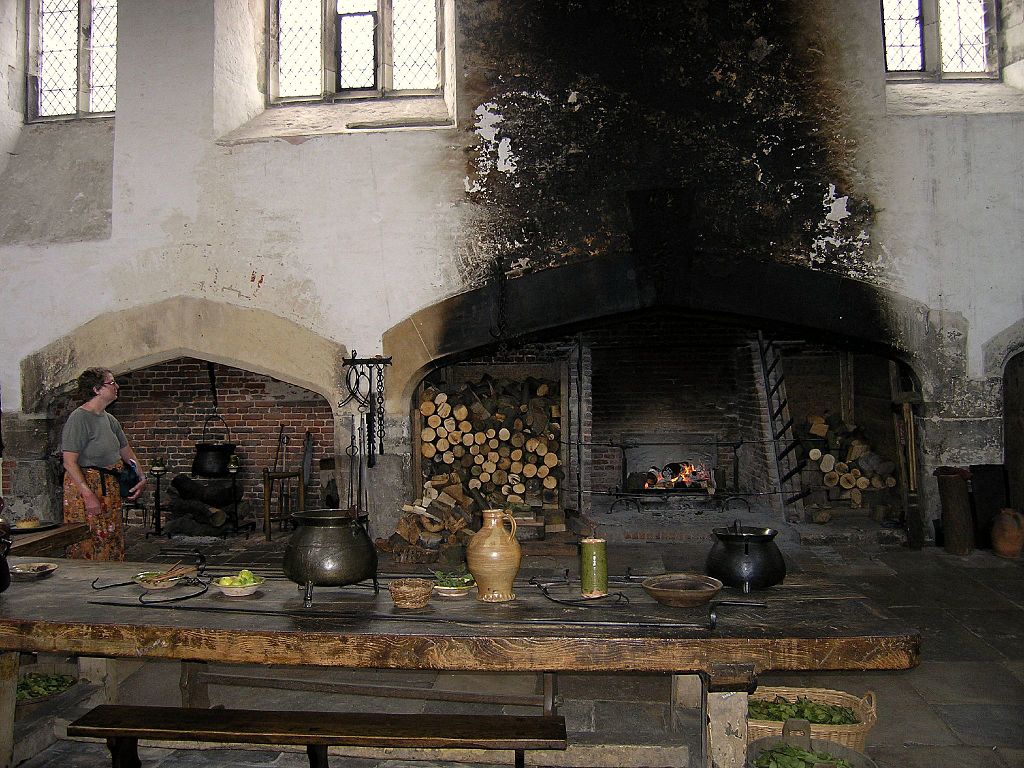 Las cocinas en Hampton Court exhiben ventanas altas en la pared blanca, una enorme chimenea con asadores y una pared ennegrecida arriba. Mesas con herramientas al frente