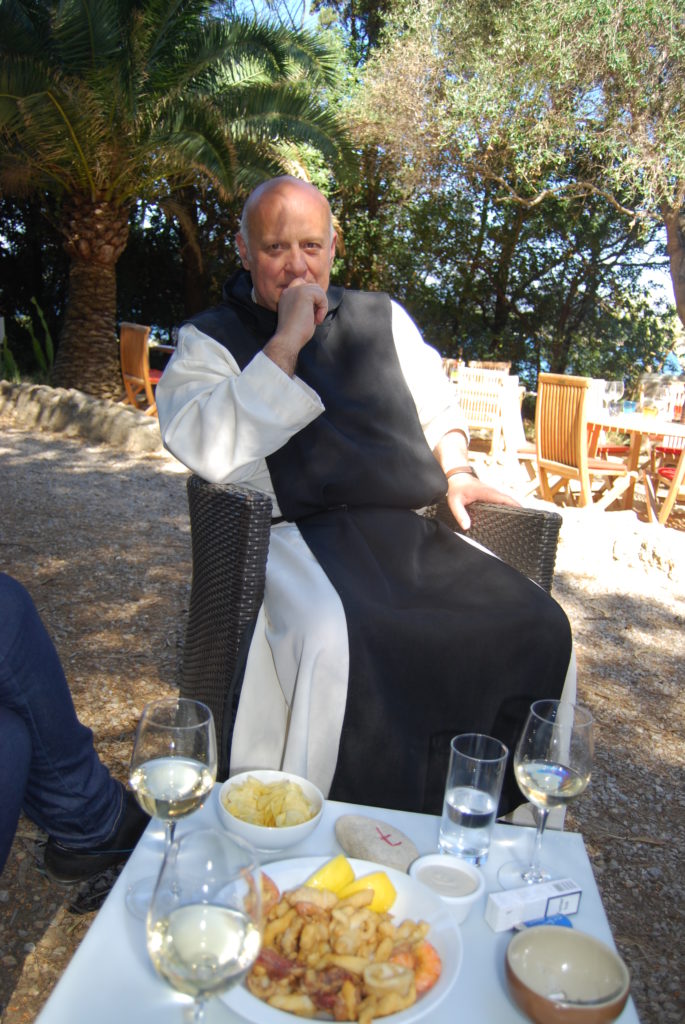 Un monje en una silla al aire libre con una mesa de comida y vino frente a él en Naomh Onórai