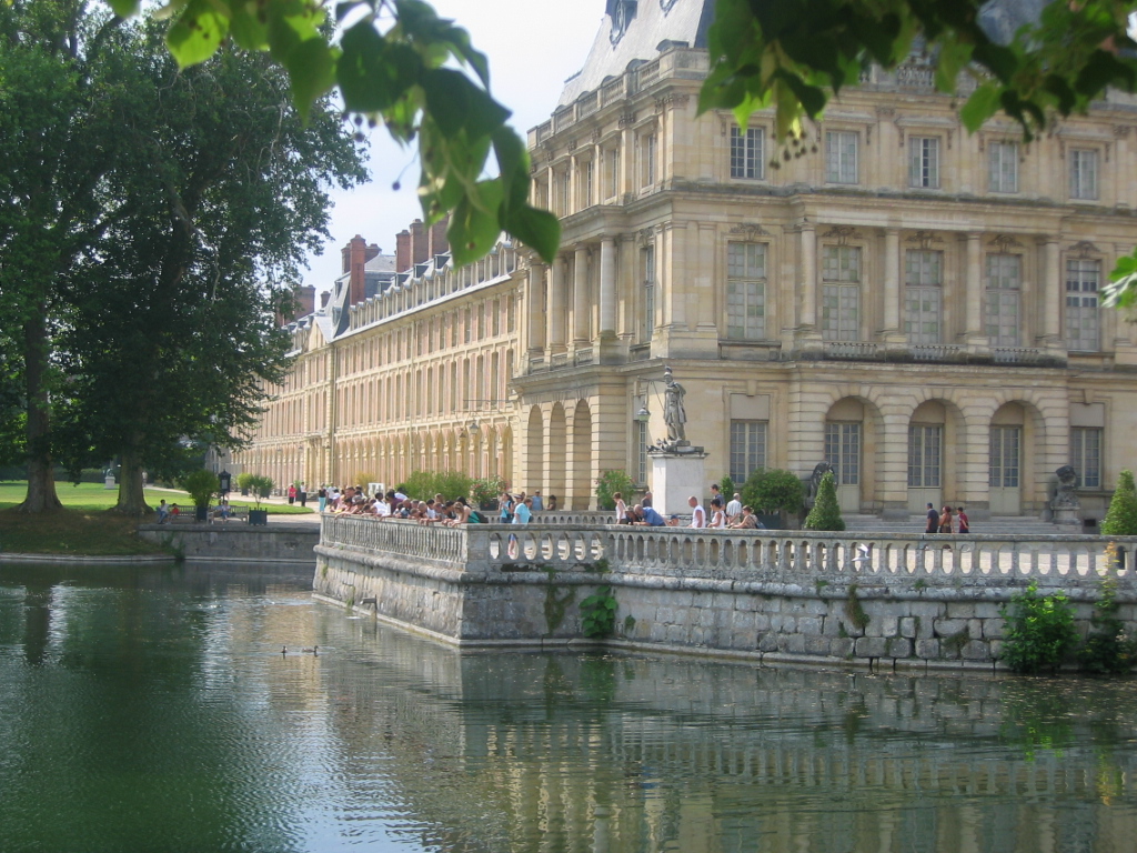 Vista exterior del castillo de Fontainebleau. Vista de esquina desde el lago con balaustrada y gente frente al edificio de piedra suave de 4 pisos