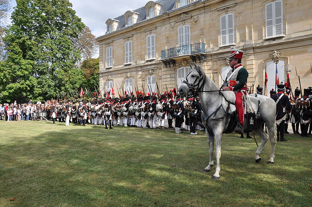 Napoleón en París Château de Bois-Préau con personas vestidas como soldados de Napoleón y un hombre a caballo frente a un bonito edificio neoclásico de 3 pisos en el césped