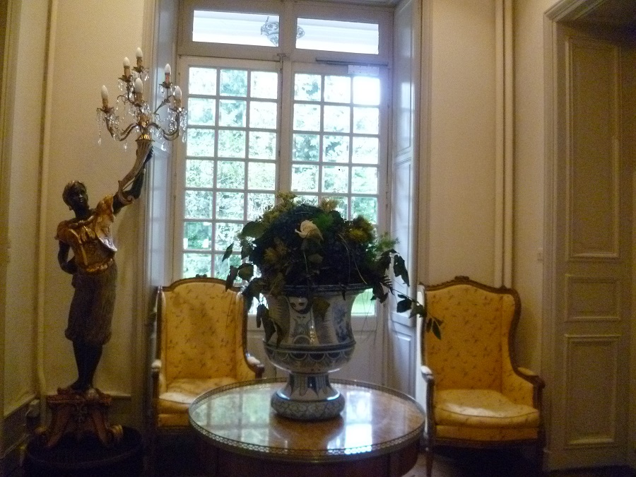 La casa de Napoleón en Ile d'Aix que muestra una habitación modesta con una ventana detrás, sillas, una mesa con flores y una figura negra elaborada que sostiene un candelabro para iluminarse.