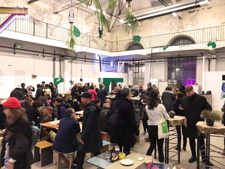 La FABuleuse Cantine en la Bienal de Diseño que muestra un edificio industrial con un gran espacio hasta el techo, mesas, sillas y muchos jóvenes comiendo