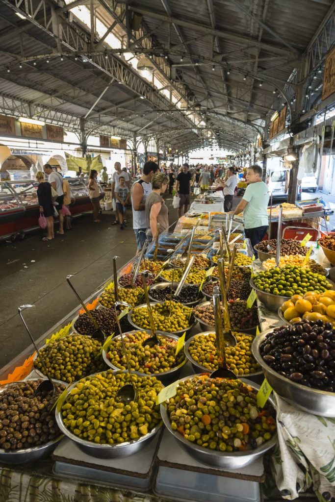 Muchas aceitunas diferentes apiladas en el puesto de aceitunas del mercado de Antibes con gente en el fondo