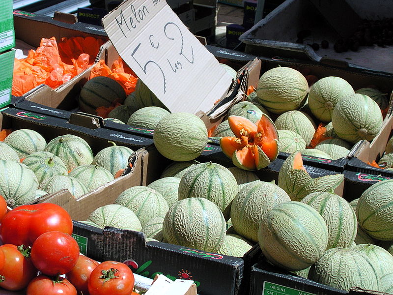 Melones maduros en una mesa con tomates y un cartel de precio