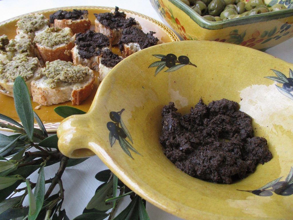 Cinta hecha de aceitunas negras en un recipiente amarillo con rebanadas de pan francés y tapenade en la parte posterior 