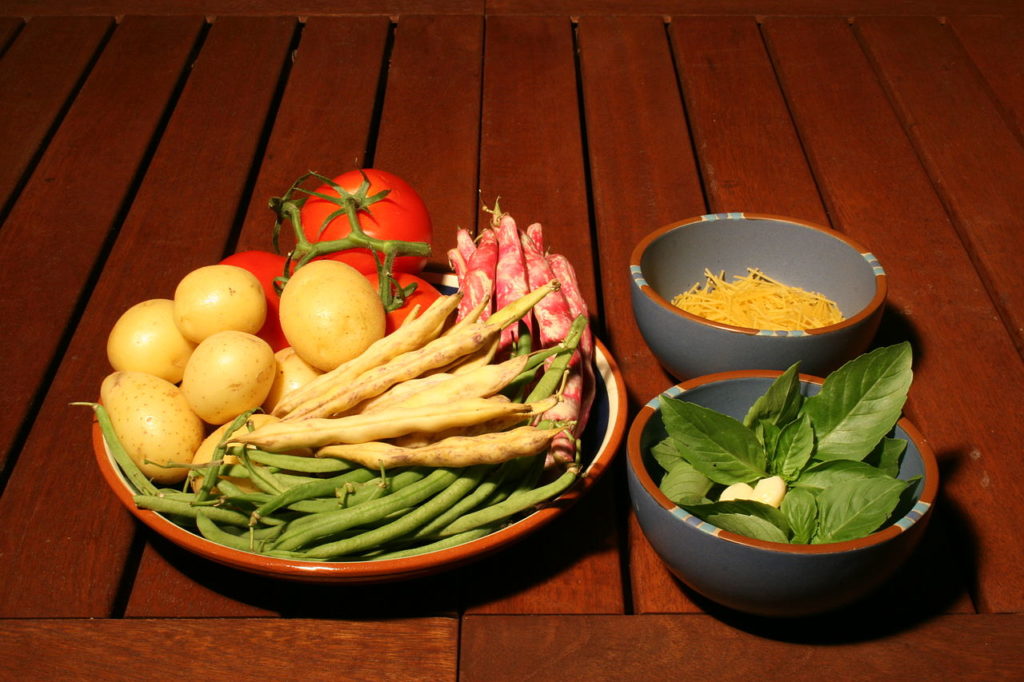 Ingredientes de pistou en tres tazones con verduras frescas en uno, hojas de albahaca y piñones en el segundo y queso en uno en la parte de atrás sobre una mesa con listones de madera