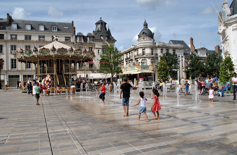 Ponga Martroi en Orleans mostrando una gran plaza con fuentes que brotan del suelo, niños y familias jugando; rotonda a la izquierda y edificios antiguos detrás