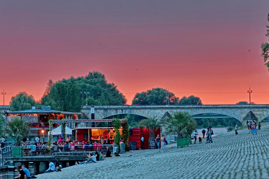 Café La Sardine a orillas del río Loira Orleans al amanecer con un puente al fondo, árboles y mesas y sillas al aire libre