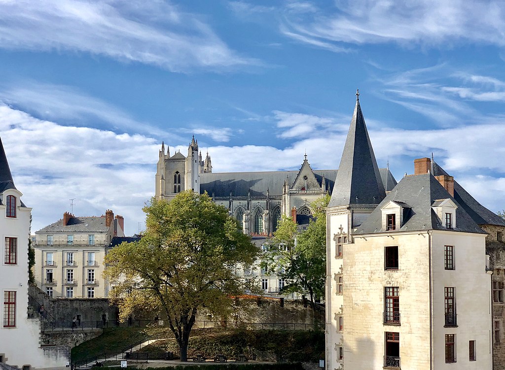 La ciudad de Nantes que incluye un edificio alto y una torre de palacio, con una catedral y otros edificios clásicos al fondo y una gran nube de cielo azul