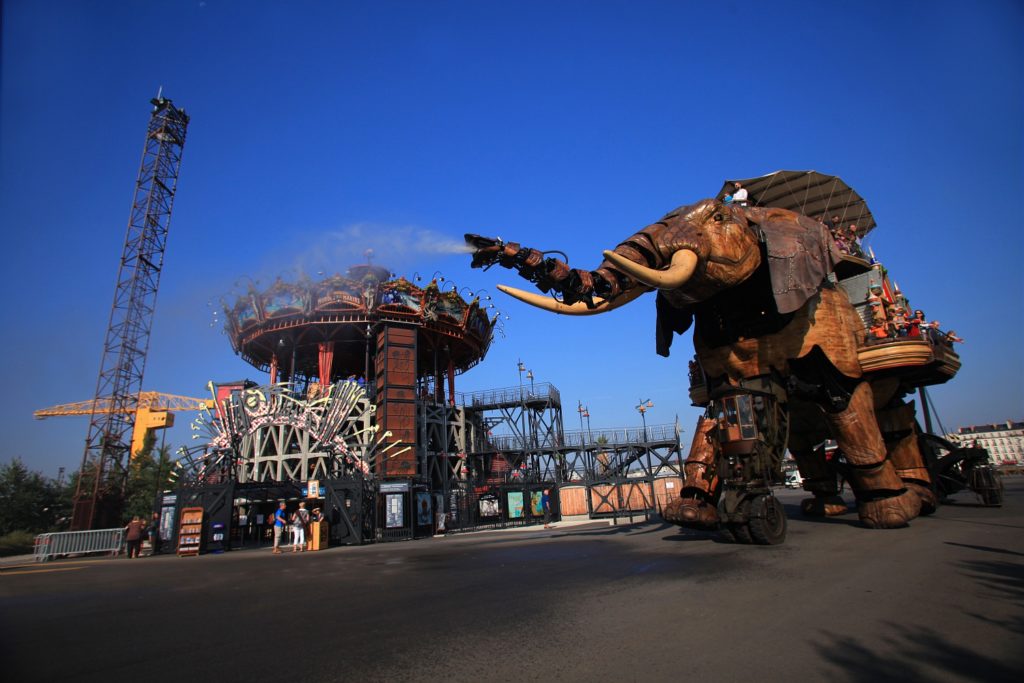 El Elefante Mecánico de Nantes con gente a lomos echando agua hacia otra máquina carrusel