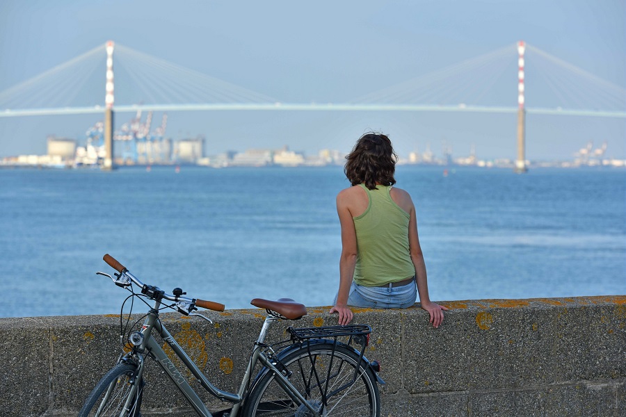 Detrás de la niña sentada en un banco verde con una bicicleta detrás de ella mirando el mar azul hasta el enorme puente de St Nazaire y la ciudad más allá