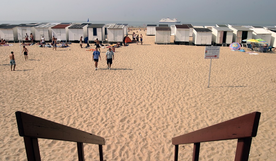 Una vista de los pasteles de playa en Calais plage con el sol al frente, cabañas de playa detrás y gente en el medio