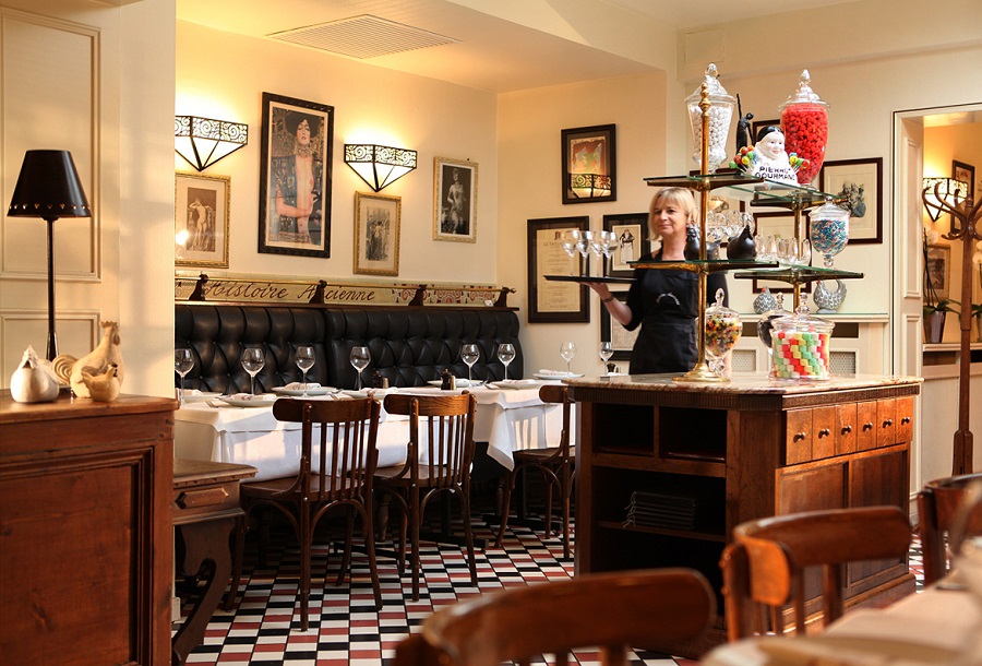 Interior del bistró Histoire Ancienne en Calais, que muestra el interior Art Deco con carteles e impresiones en paredes amarillas, mesas y sillas de madera preparadas para una comida, bar y un camarero con trav