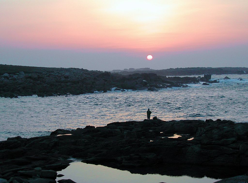 Atardecer en Ile Grande Pointe de Toual con cielo rojo, mar azul y rocas con un pequeño pescador solitario