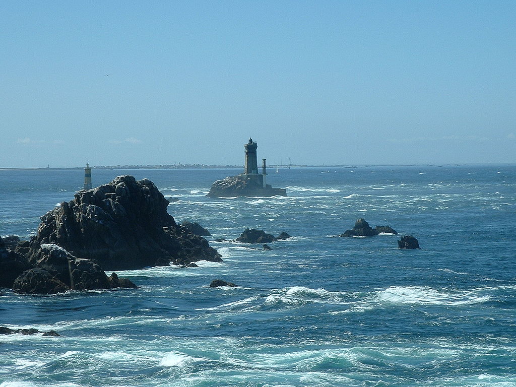 El mar embravecido en Ile de Sein en Bretaña se estrella contra las rocas en el mar con un faro en una roca en la distancia