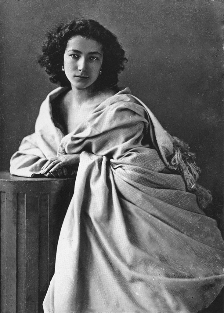 Retrato de Sarah Bernhardt por Felix Nadar en 1864. Blanco y negro apoyada en una pequeña columna con cabello largo y negro, un impresionante vestido con hombros descubiertos y una mirada ardiente.