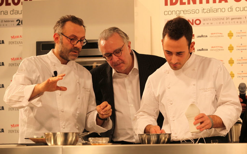 Alain Ducasse con chaqueta y camisa entre 2 jóvenes chefs viéndolos preparar la comida