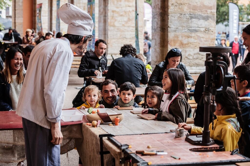 Un chef con un toque enorme delante de las mesas y los niños detrás a la altura de la mesa aprendiendo a cocinar