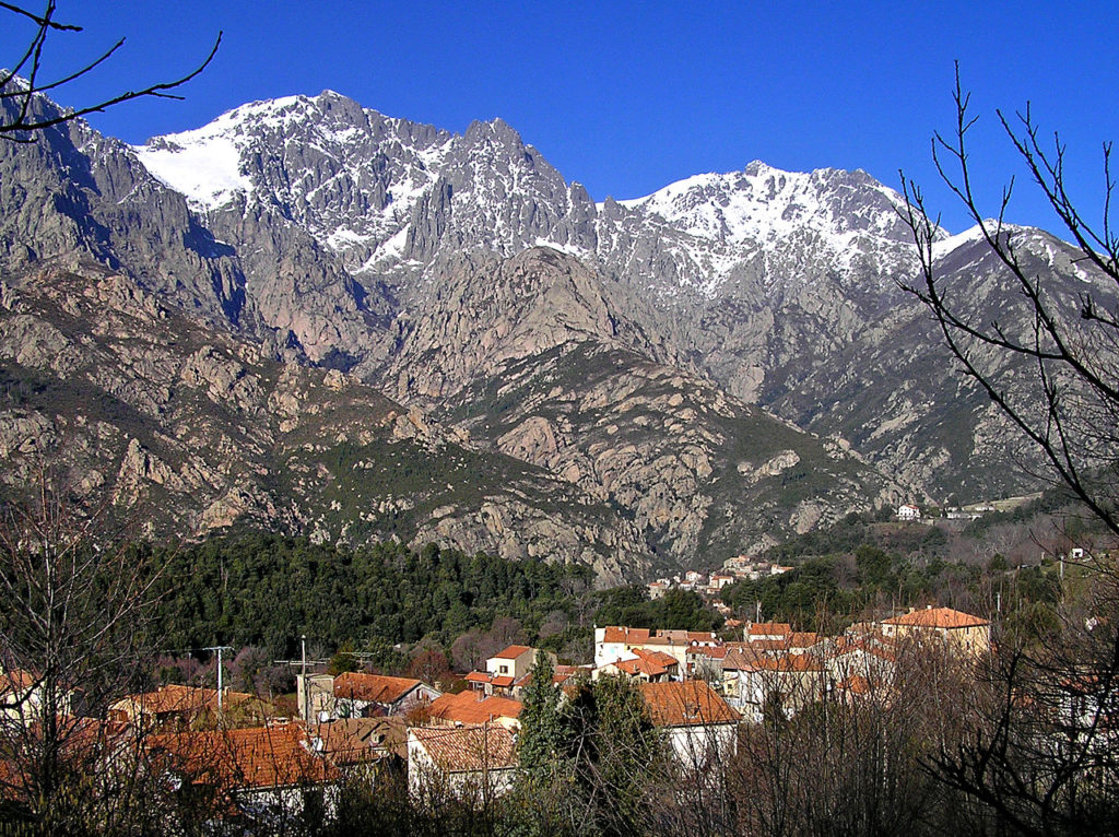 Bocognano, Córcega. Una vista larga con montañas cubiertas de nieve en el fondo y el pequeño pueblo en un valle debajo rodeado de árboles. Tejados rojos y casas antiguas.