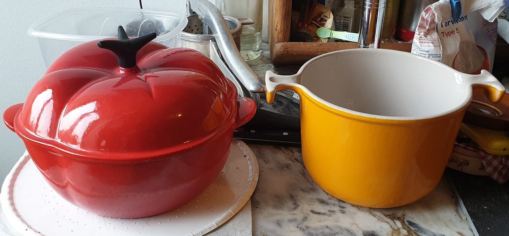 Utensilios de cocina Le Creuset con una cacerola roja en forma de tomate a la izquierda y una olla naranja abierta sin tapa a la derecha