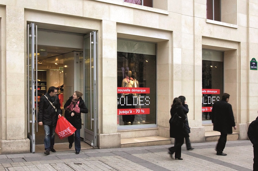 Escaparate con escaparates con letreros de Soldes y dos personas saliendo con bolsas rojas, en París