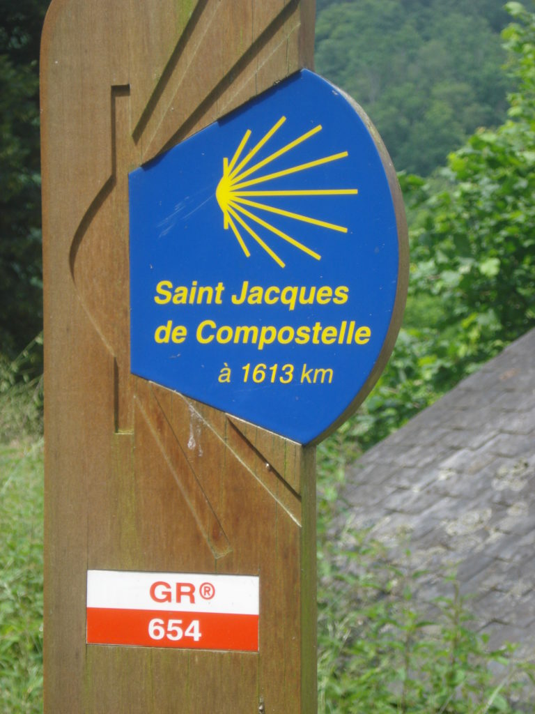 Signo público de San Jacques de Compostela. Fondo azul, una concha marina idealizada en un poste de madera con un camino detrás