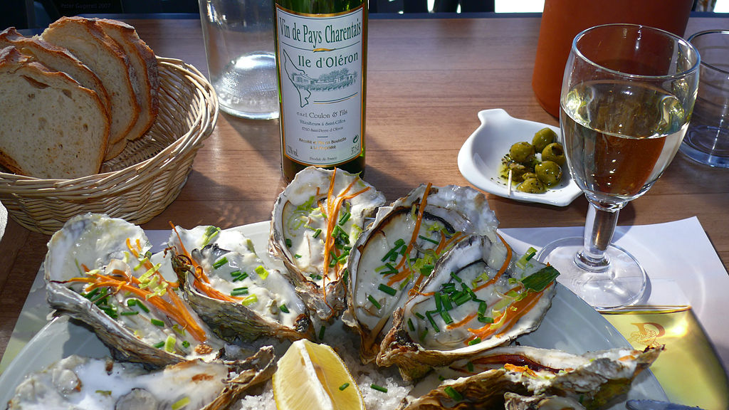 Elija una comida de ostras en conchas con perejil verde y limón al frente con una botella de vino Oleron en la parte posterior y otros platos pequeños.