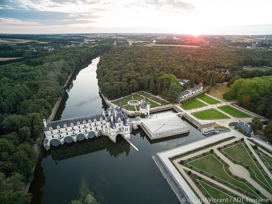 Vista aérea de Chenonceau que muestra un edificio blanco al otro lado del río con jardines formales a la derecha dispuestos en patrones geométricos y una vista que se extiende a lo lejos