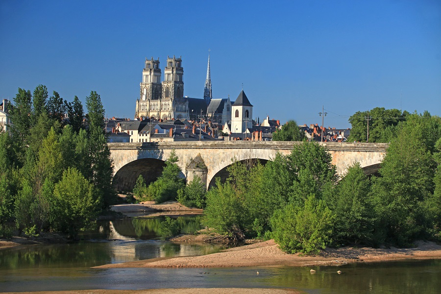 Catedral de Sainte Croix en Orleans desde el otro lado del río Loira con torres altas en el frente, un puente y una pequeña isla con árboles.