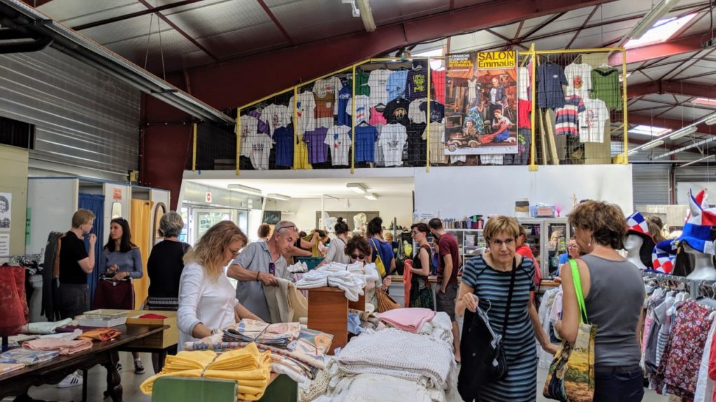 Emaús en Le Puy-en-Velay. Gran espacio industrial en el interior con personas revisando montones de cosas extrañas en las mesas y más artículos apilados en el espacio del techo