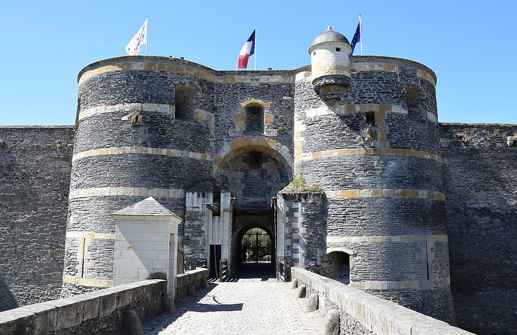 Entrada al Chateau d'Angers que muestra un largo camino de grava que conduce a la puerta que llena una imagen con dos enormes torres redondas en paredes de piedra negra con líneas de piedra blanca