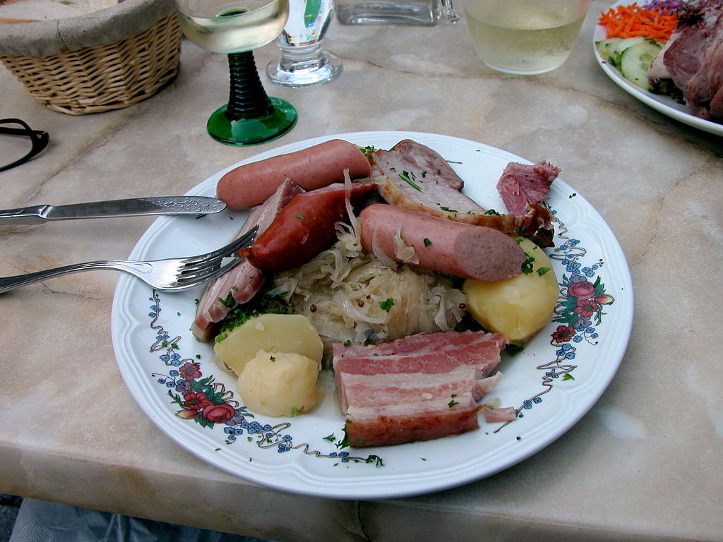 Alsation francés Choucroute plato de comida con un plato lleno de jamón, patatas, salchichas y verduras, con una copa de vino en el fondo