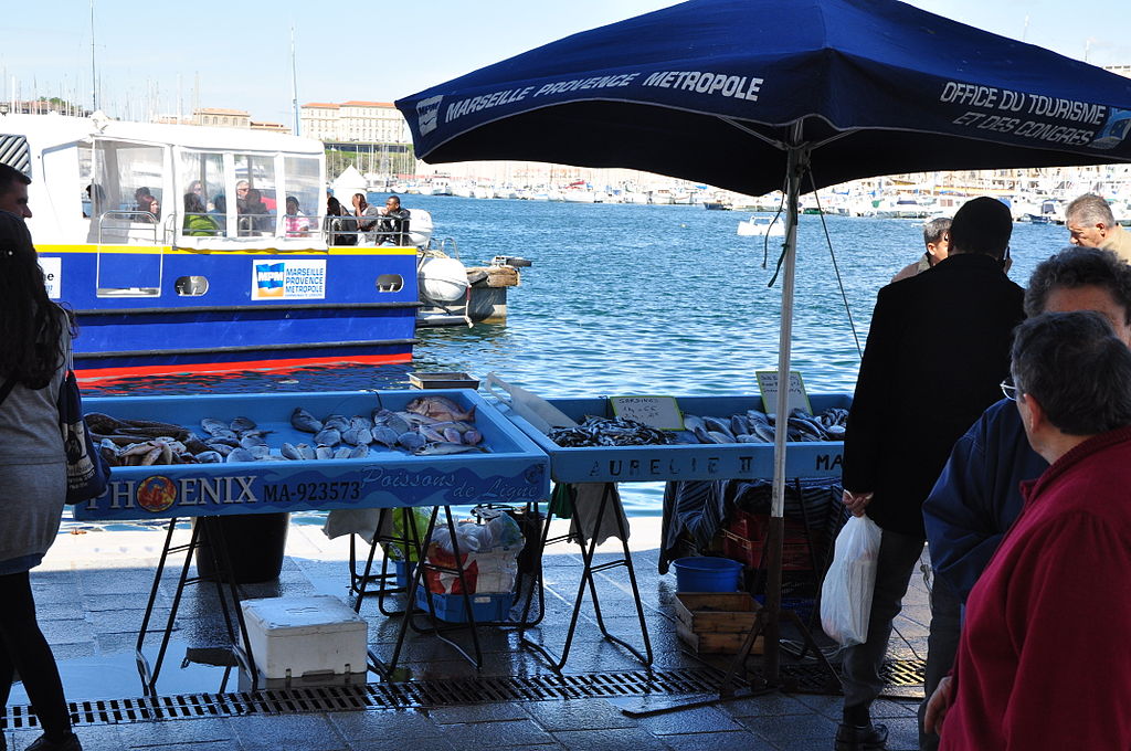 Mercado de pescado de Marsella con un puesto y una sombra azul con pescado frente al agua con un ferry detrás y casas