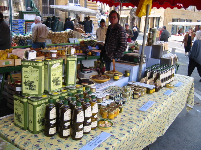 Un puesto en el mercado de Avignon con un titular de puesto en la parte trasera y una mesa con un mantel Probencal lleno de botellas y latas de aceite de oliva