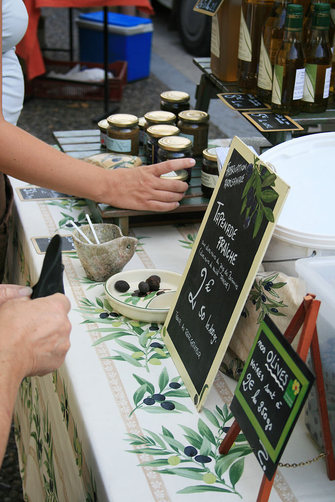 Mercado provenzal Vaison la romaine con puesto de tapenade, mantel decorado con aceitunas y tarros de tapenade con brazo extendido