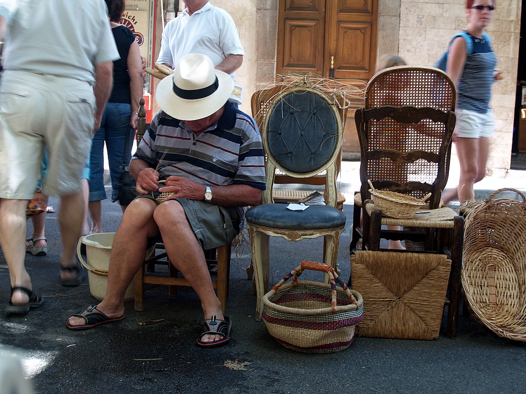 Mercado de Alernes Provence con un hombre en un panamá blanco en una silla mirando hacia abajo mientras enlata un asiento y otras sillas a su lado